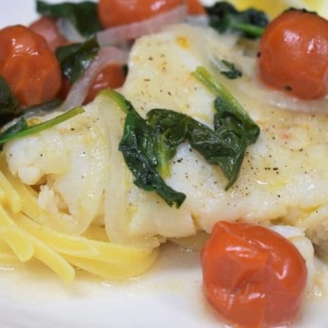 A close up picture of cod, tomatoes and spinach served over pasta on a white plate.