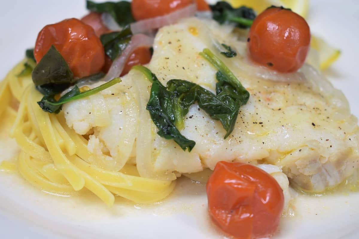 A close up picture of cod, tomatoes and spinach served over pasta on a white plate.