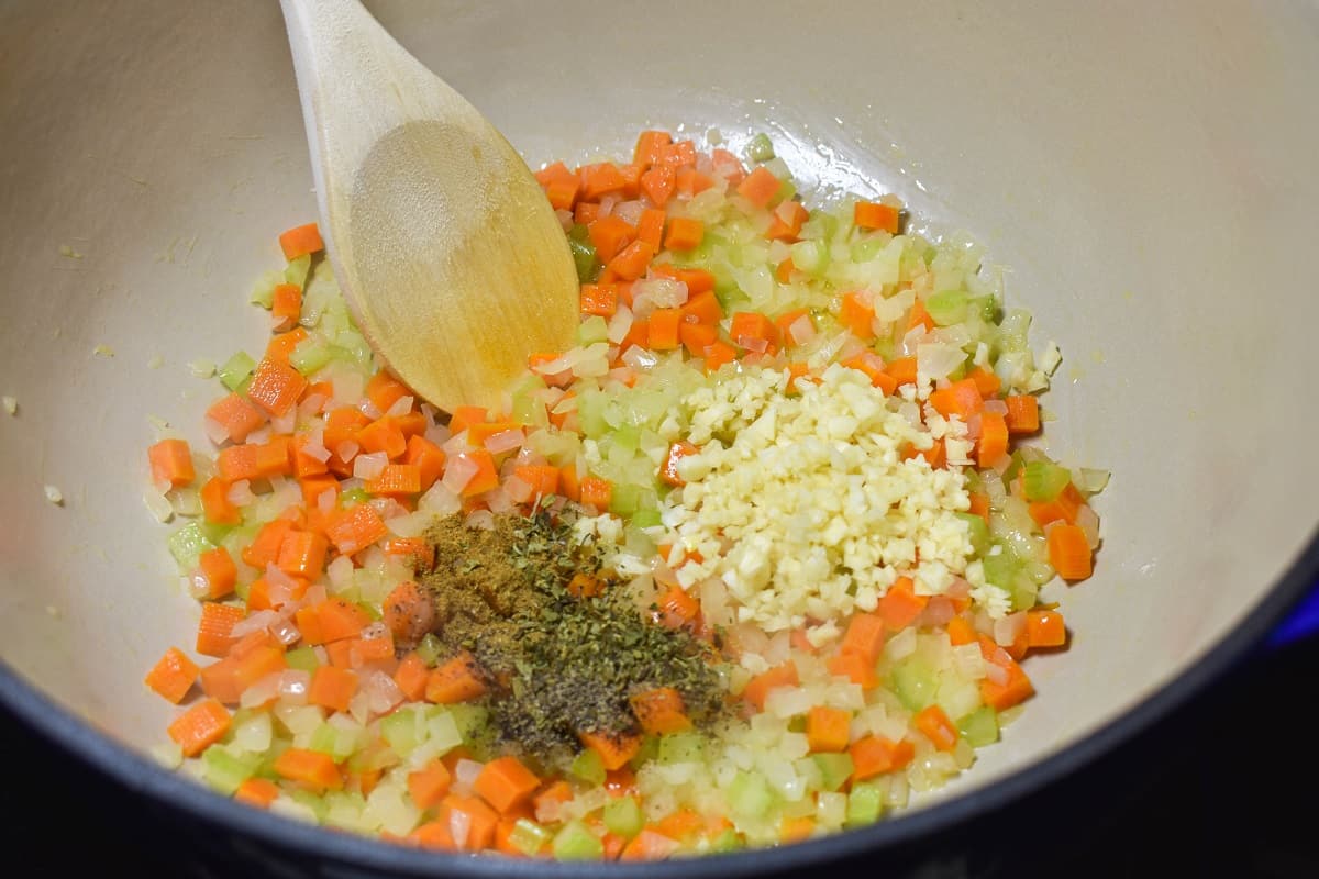Onions, carrots, celery, minced garlic and spices sauteing in a large pot with a wood spoon in the pot.
