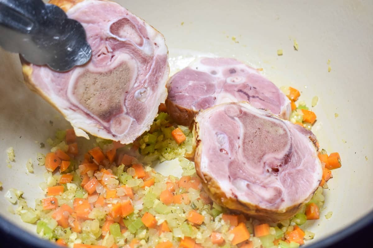 Three slices of ham shank being added to the vegetables cooking in the pot.