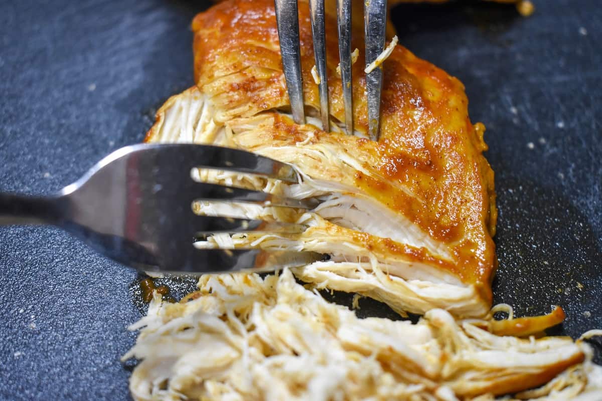 Two forks shredding a cooked chicken breast on a black cutting board.