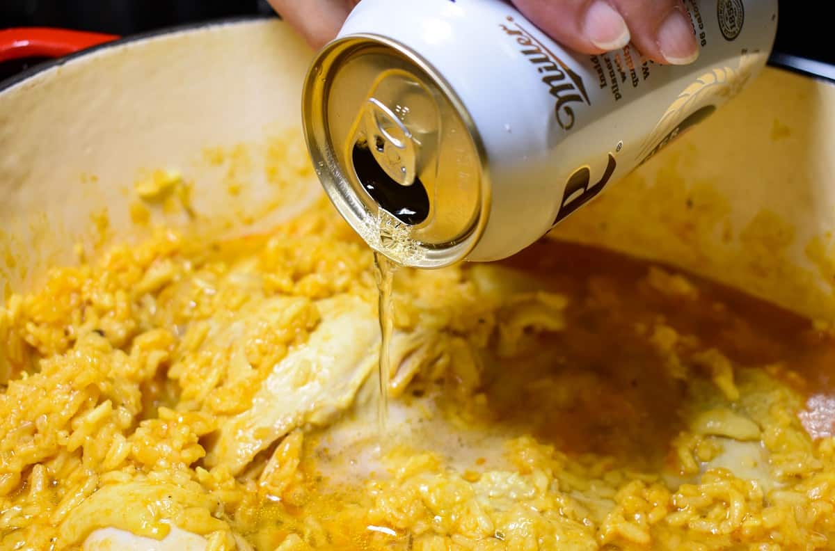A beer being added to arroz con pollo in a pot