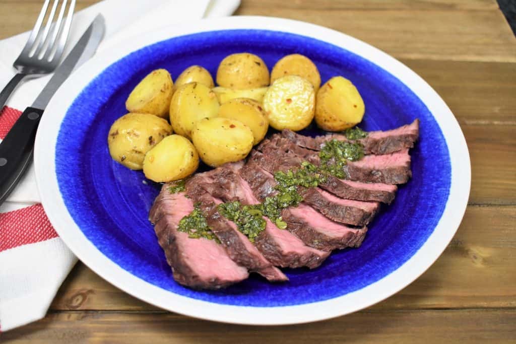 Grilled potatoes with sliced flat iron steak topped with chimichurri and served on a blue plate with a white rim.