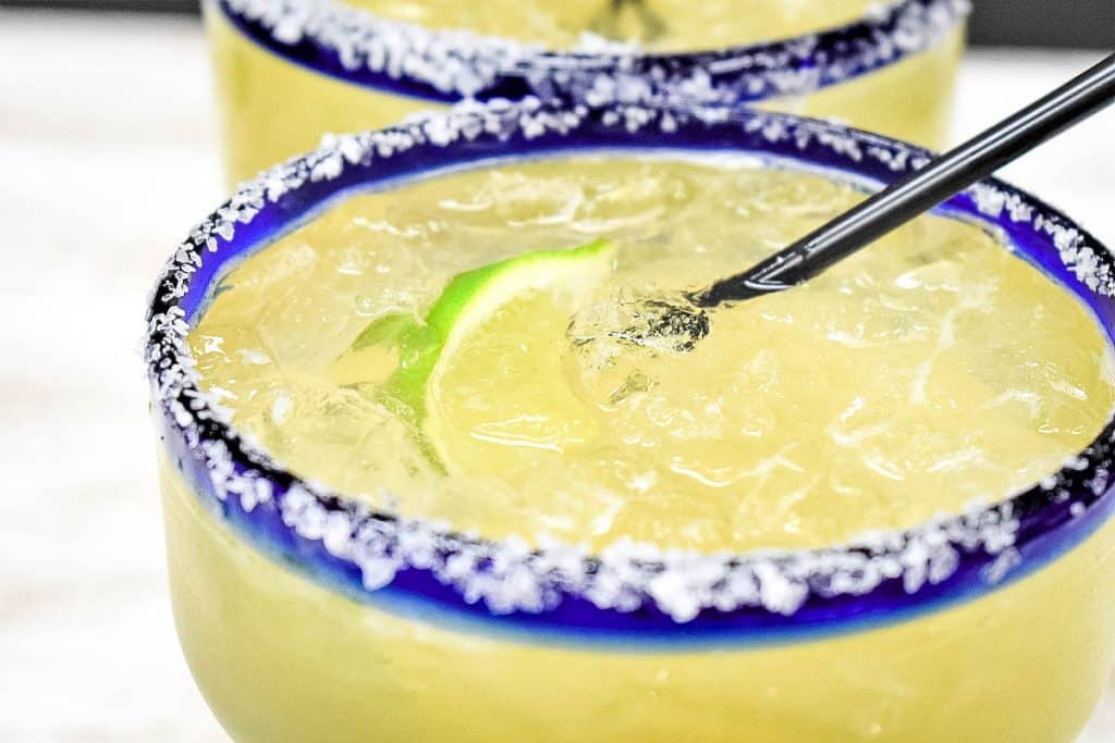 A close up picture of a margarita with a blue, salted rim, a lime wedge floating in it with a black straw.