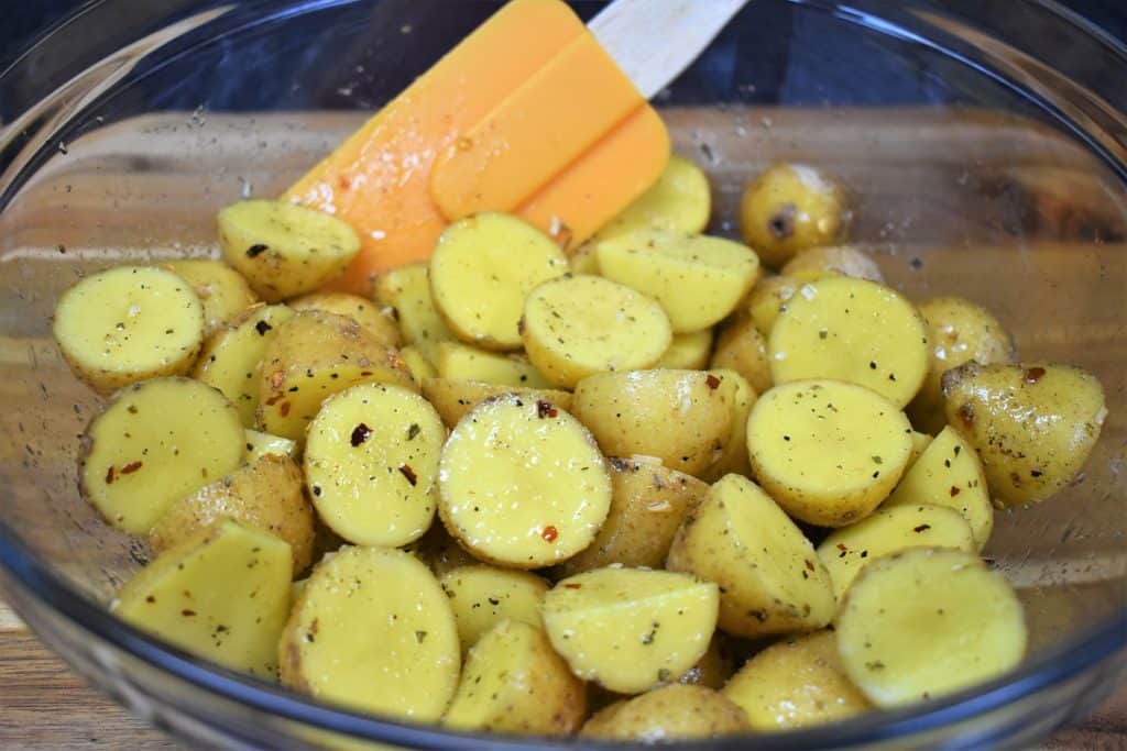 Raw, halved honey gold potatoes tossed with oil and seasoning mix in a large clear glass bowl.
