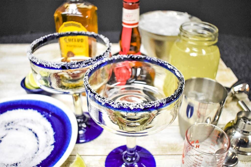 Ingredients for a margarita displayed, with an empty margarita glass with a salt rim in the front.
