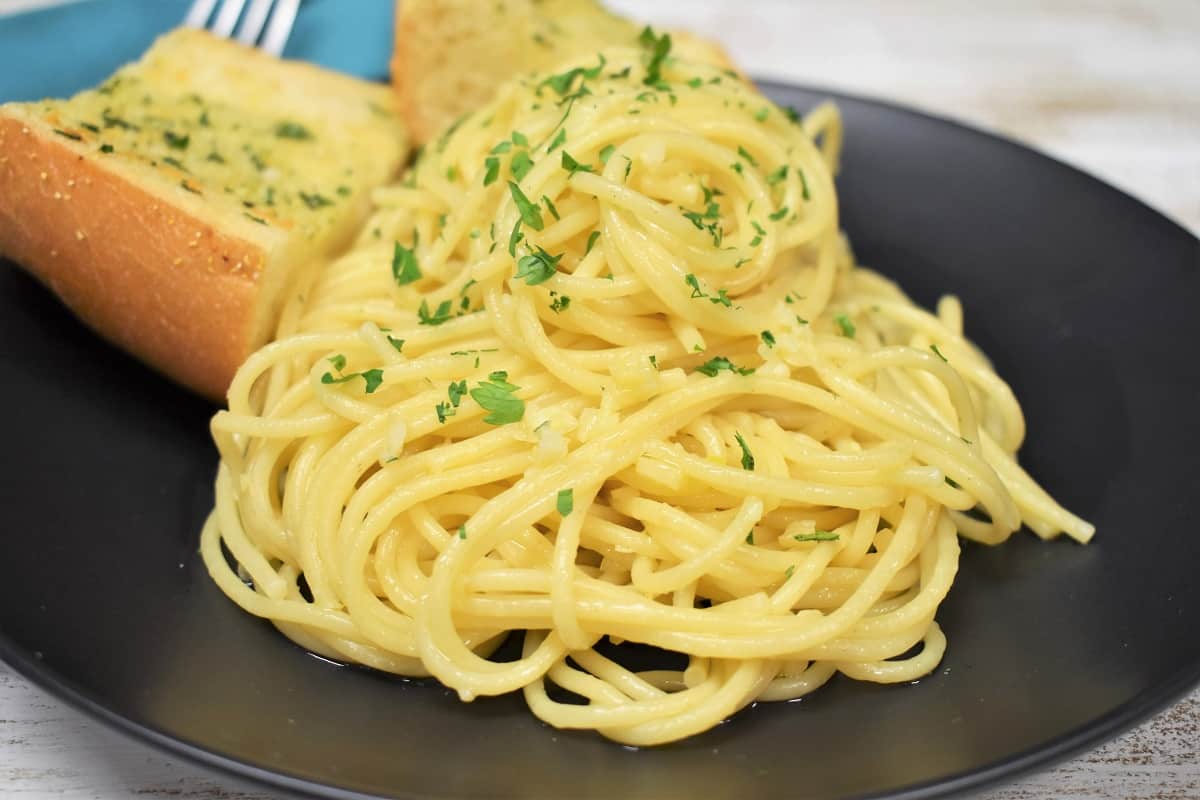 A close up of spaghetti aglio e olio served on a black plate of two pieces of garlic toast in the background.