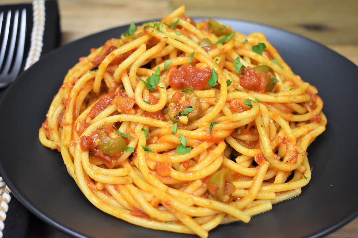 Spaghetti and a tomato olive sauce served on a black plate.