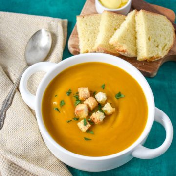 The creamy vegetable soup served in a white bowl, set on a green table with a beige linen and sliced bread in the background.