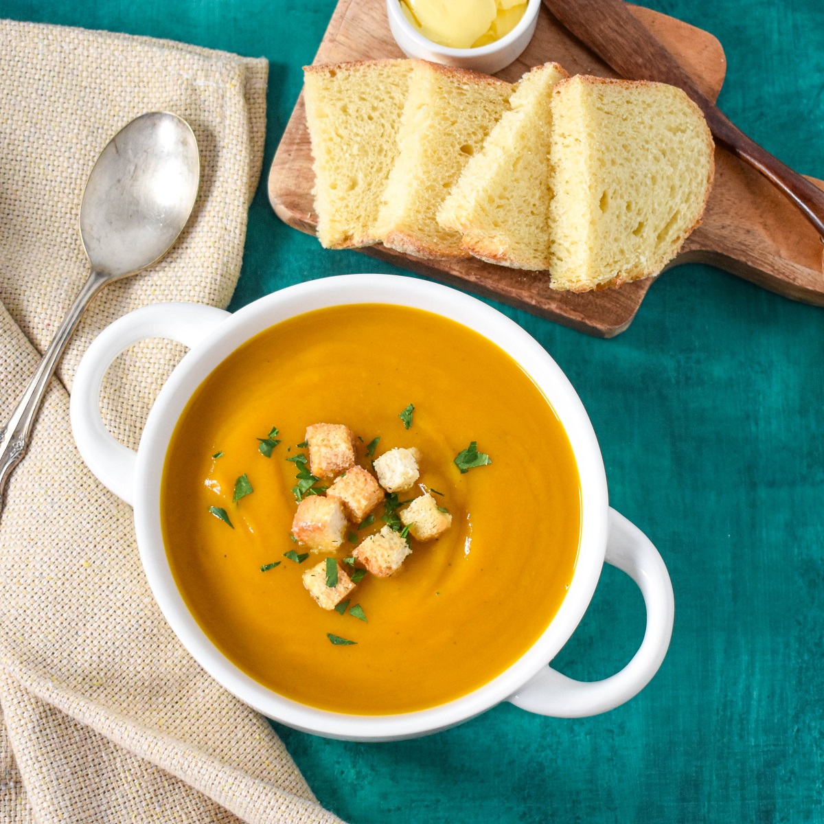 The creamy vegetable soup served in a white bowl, set on a green table with a beige linen and sliced bread in the background.