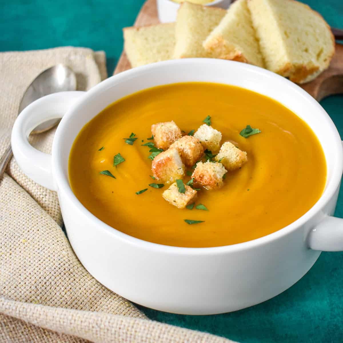 A close-up of the vegetable soup served in a white bowl with a beige linen and bread slices in the background.