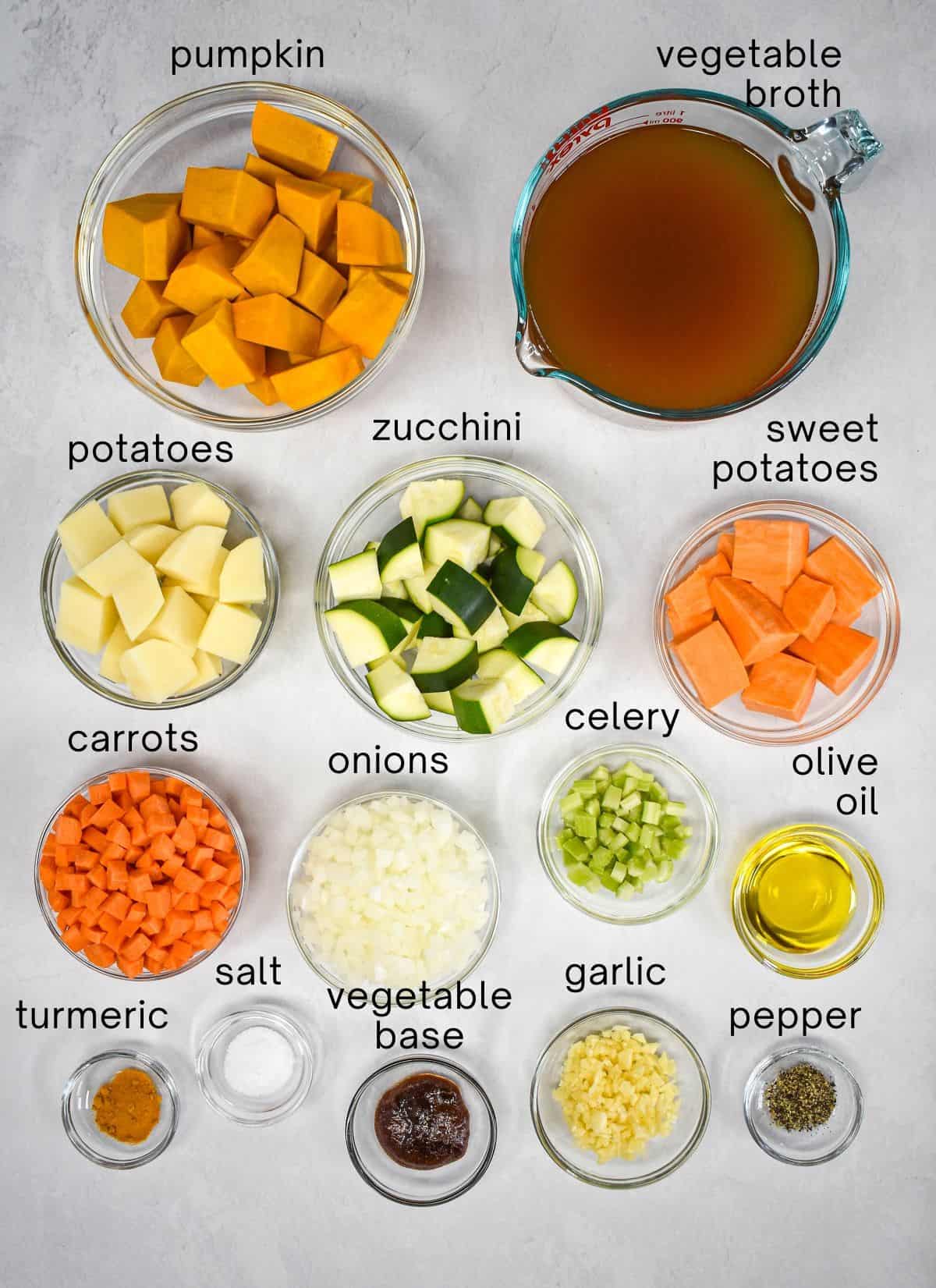 The ingredients for the vegetable soup prepped and arranged in glass bowls set on a white table with each labeled with small, black letters.