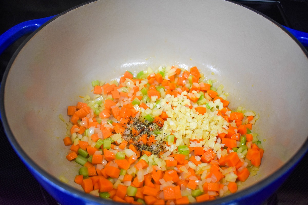 Minced garlic and black pepper added to the carrots, onions, and celery cooking in a large, blue and white pot.