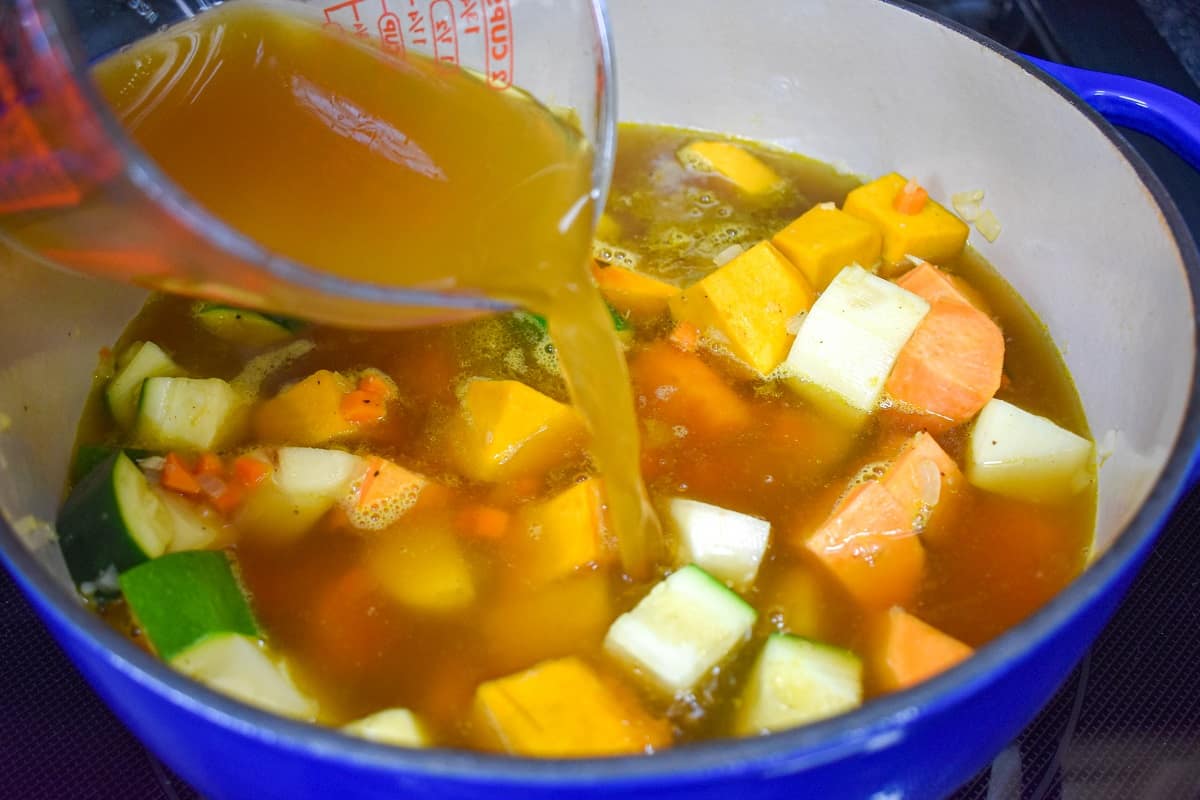 Broth being added to the array of cut vegetables in a large, blue and white pot.
