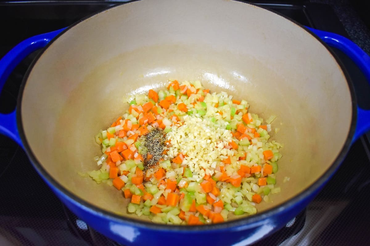 Minced garlic and pepper added to onions, carrots, and celery cooking in a large pot.