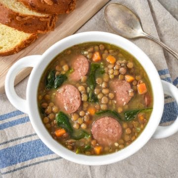 The lentil sausage soup served in a white bowl and set on a blue and beige striped linen with sliced bread in the background.