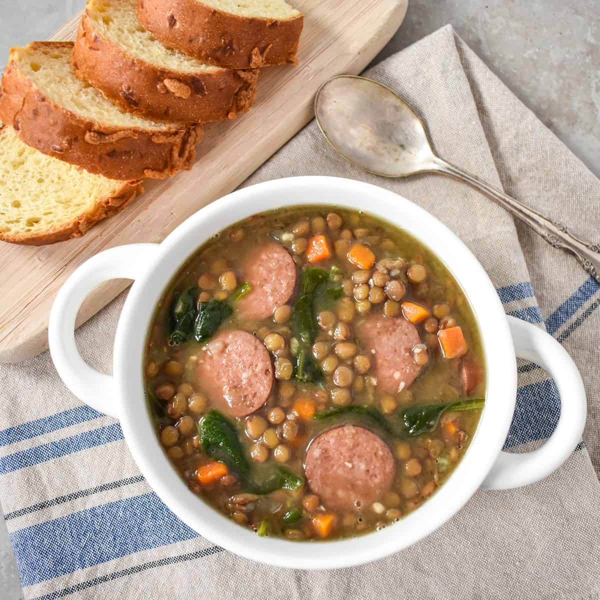 The lentil sausage soup served in a white bowl and set on a blue and beige striped linen with sliced bread on a wood cutting board in the background.