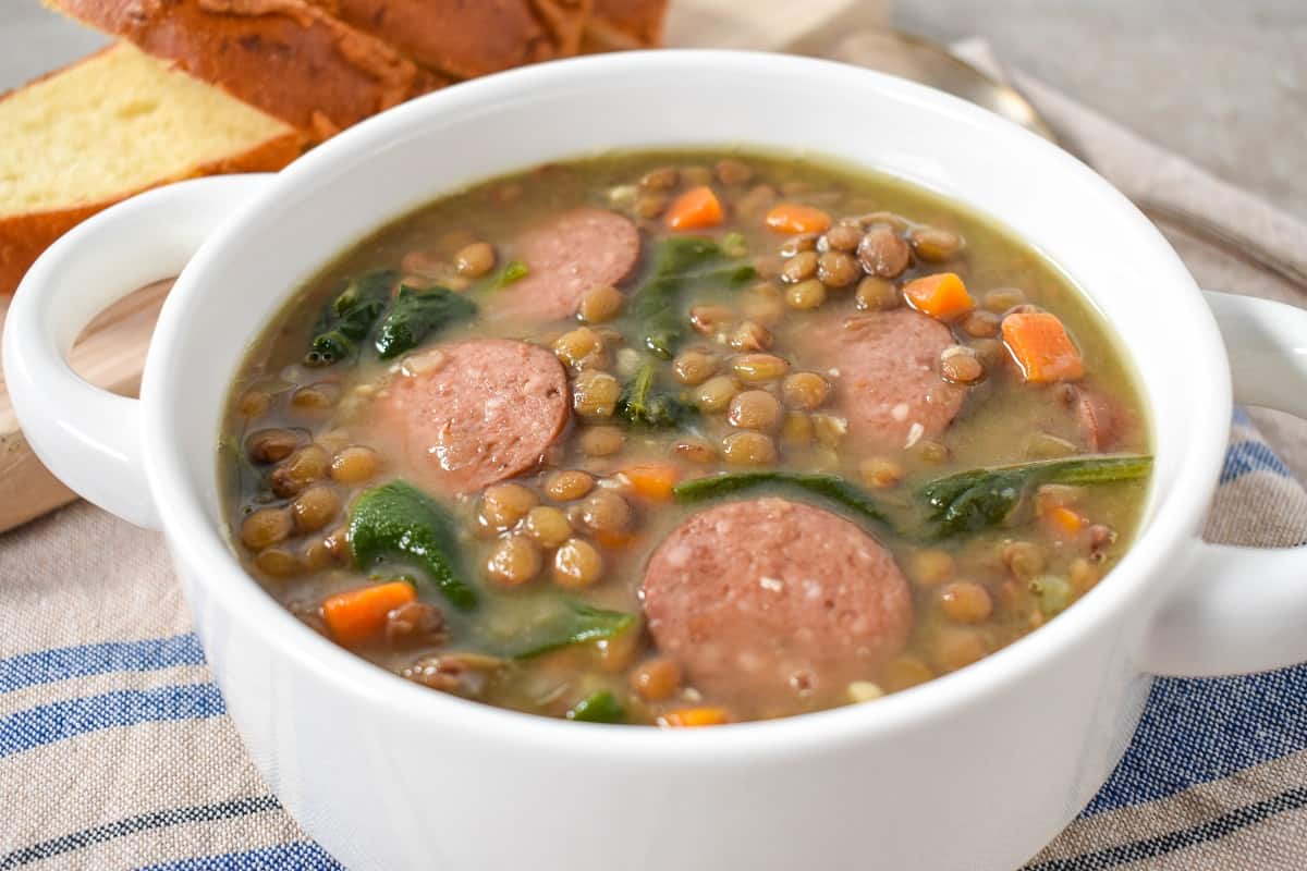 A close up of the sausage lentil soup served in a white bowl with sliced bread in the background.