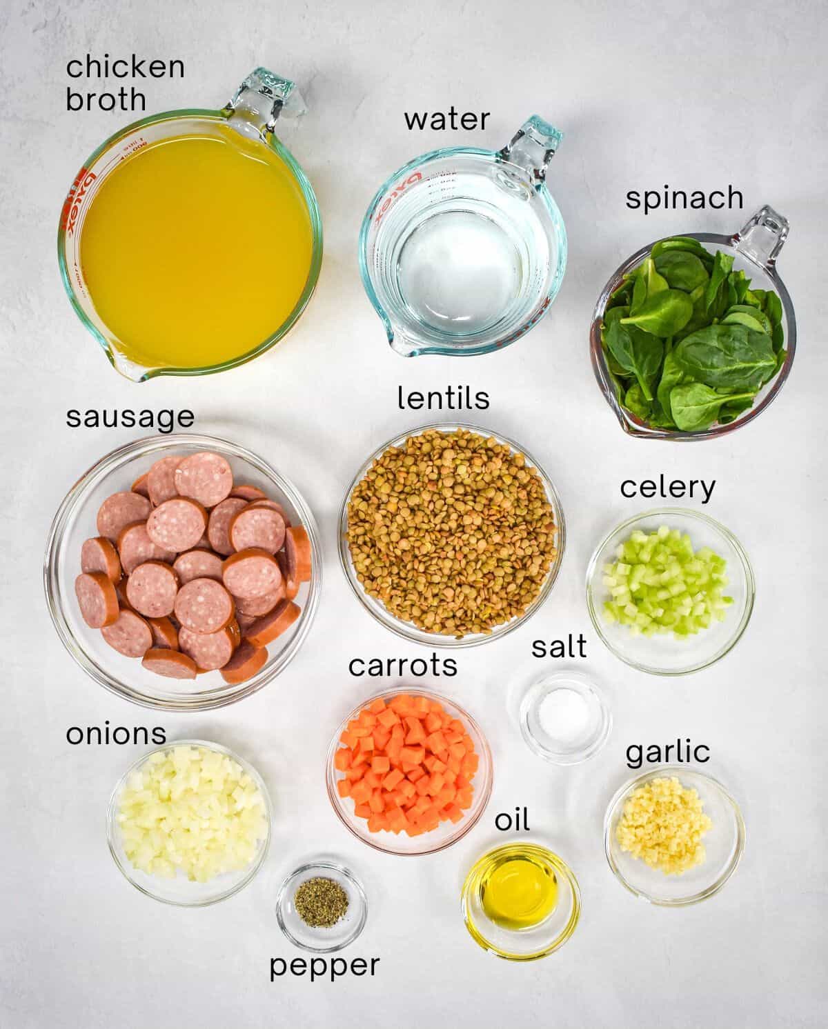 The ingredients for the lentil soup arranged in glass bowls on a white table with each ingredient labeled with small, black letters.