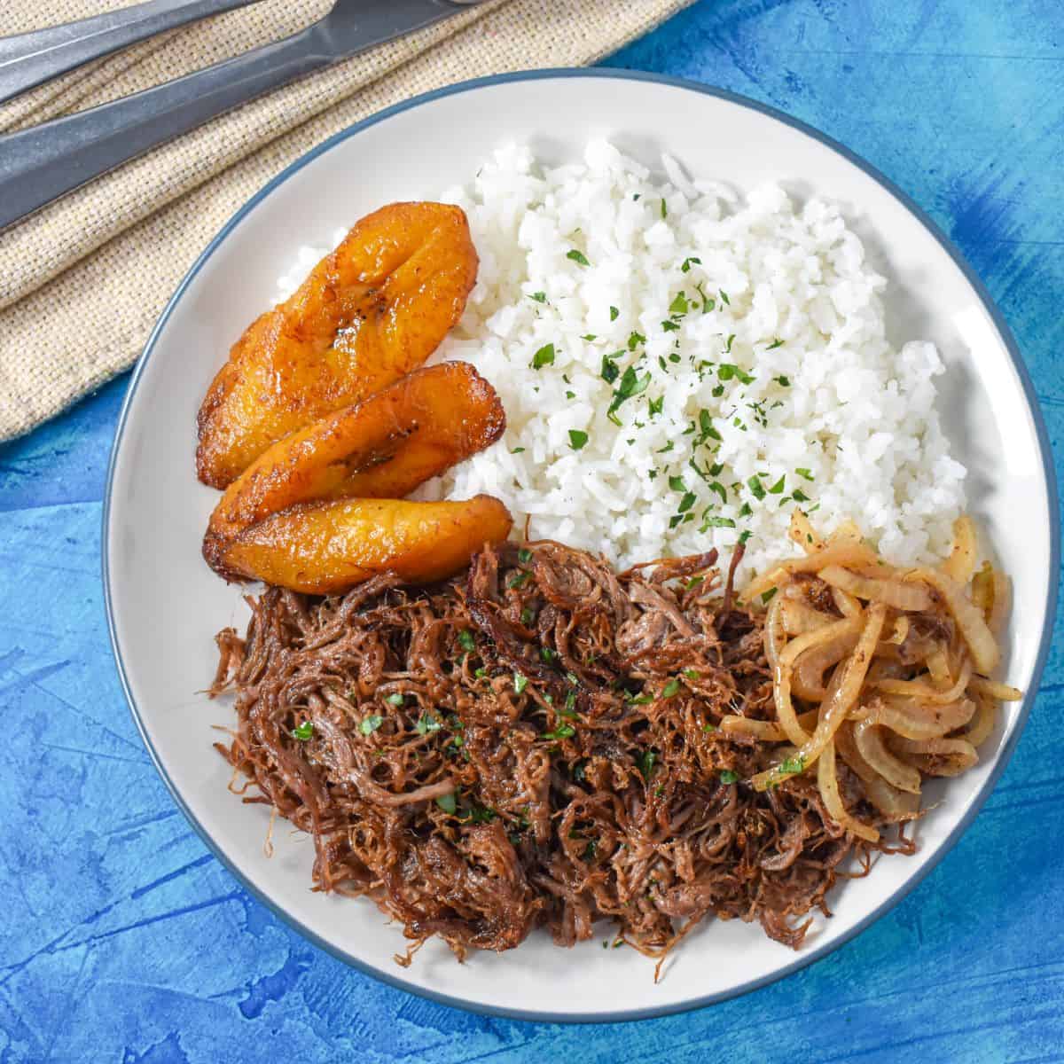 The finished fried beef served with white rice, sauteed onions and fried sweet plantains on a white plate.