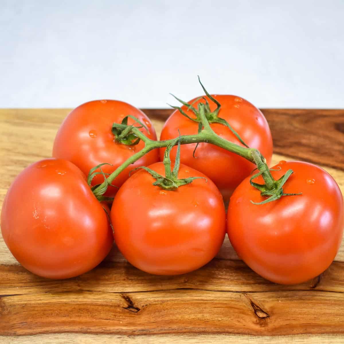 Five red tomatoes on the vine, set on a wood cutting board.