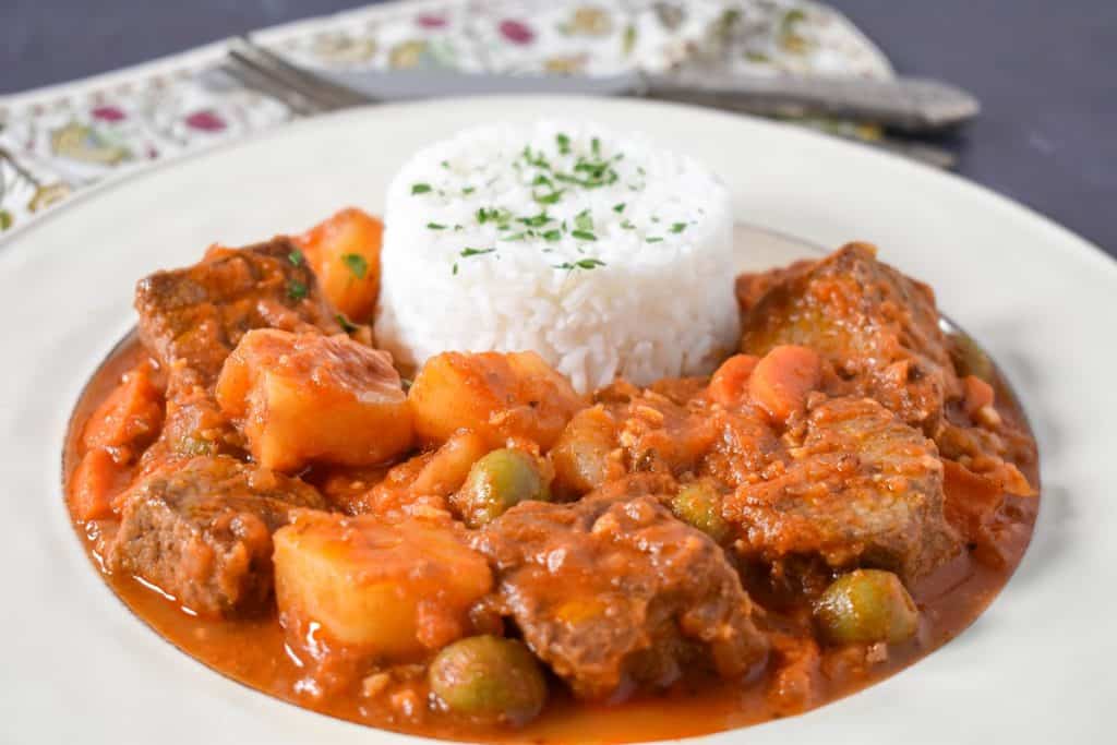 Carne con papas served with white rice in a white plate, with a linen, fork and knife in the background.