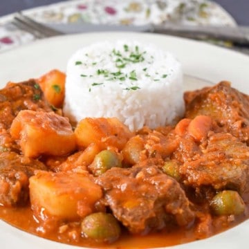 Carne con papas served with white rice in a white plate, with a linen, fork and knife in the background.