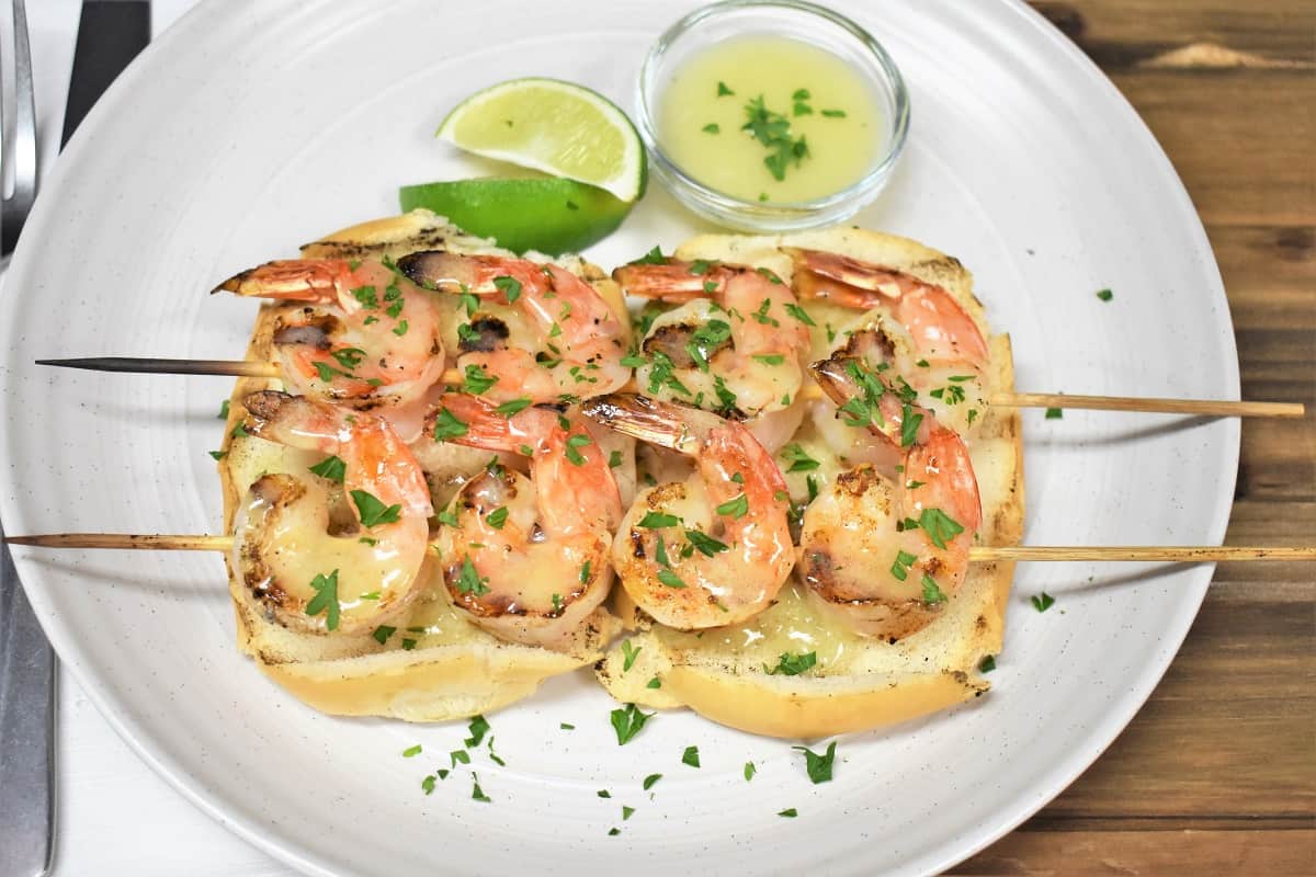 Two grilled shrimp skewers and butter lime sauce served on toasted bread on a white plate and garnished with parsley.