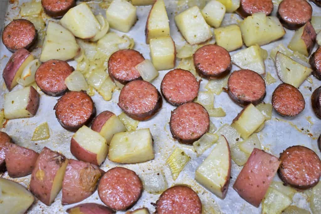 Baked sliced sausage and diced red potatoes arranged on a large baking sheet..