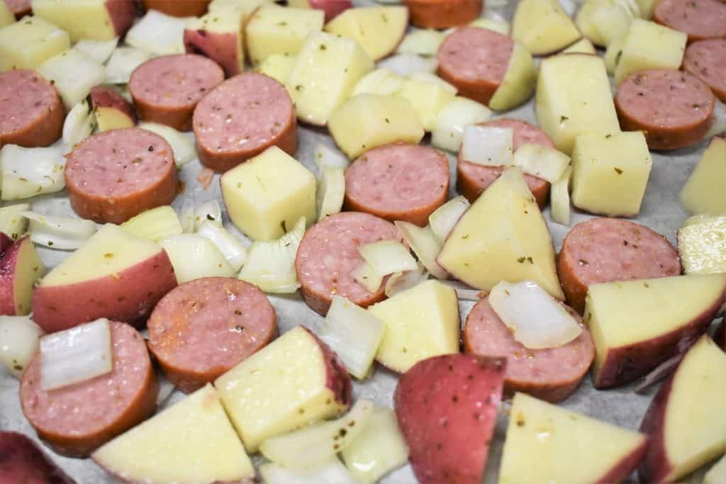 Sliced sausage, diced red potatoes and diced onions arranged on a large baking sheet, prior to baking.