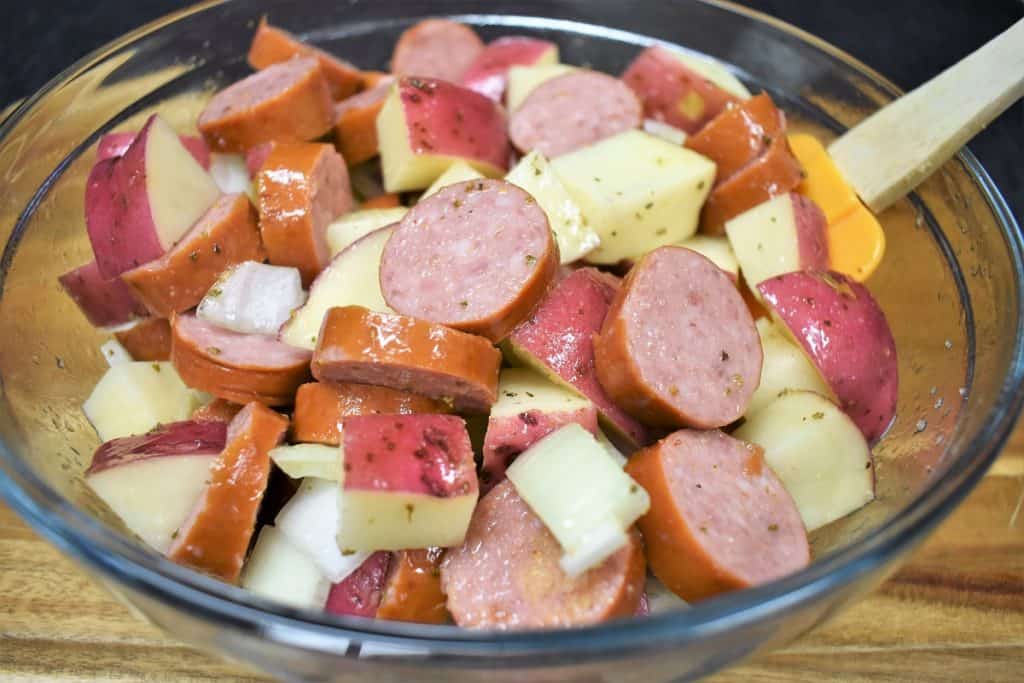 Seasoned sliced sausage, diced potatoes, and diced onions combined in a large, clear bowl.