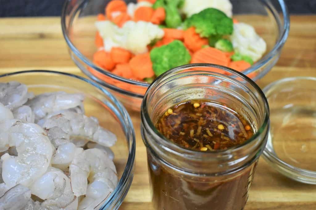 Peeled shrimp, mixed vegetables and stir fry sauce in a small jar, all displayed on a wood cutting board.