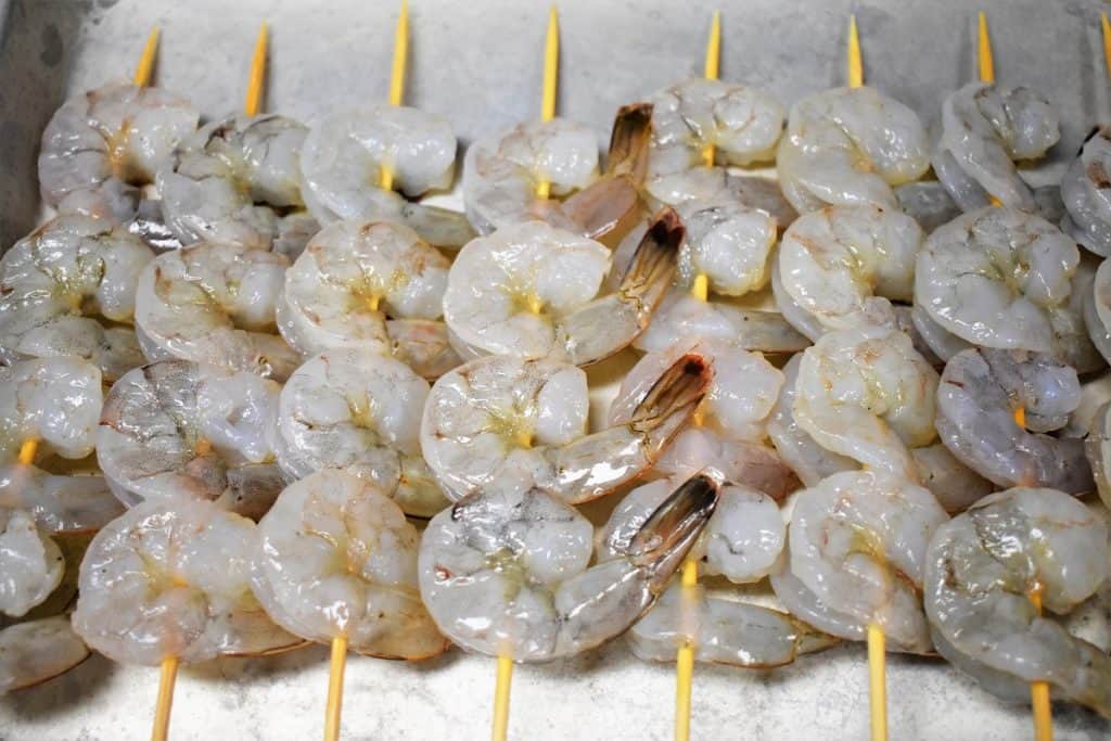 Raw shrimp on bamboo skewers arranged on a metal pan.