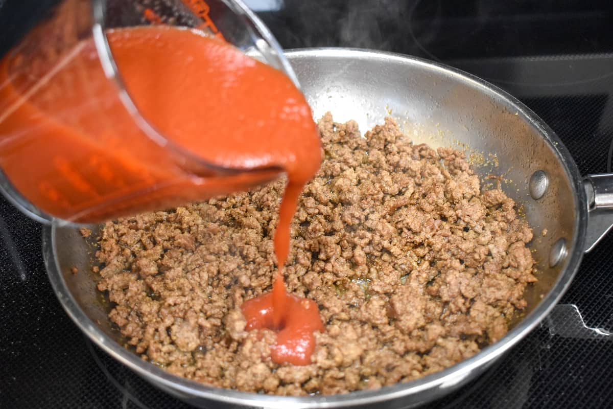 Tomato sauce being added to browned ground beef in a skillet.