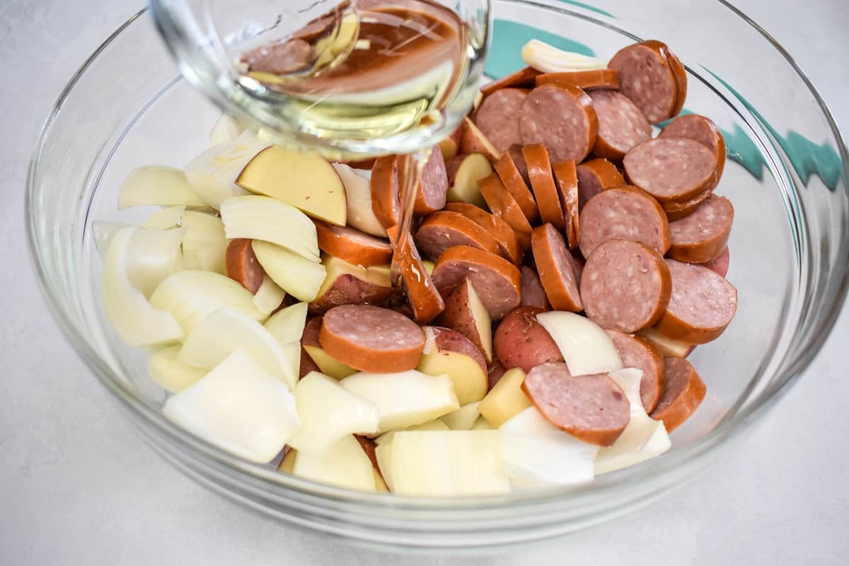 Oil being added to sliced sausage, potatoes, and onions in a large, glass bowl.