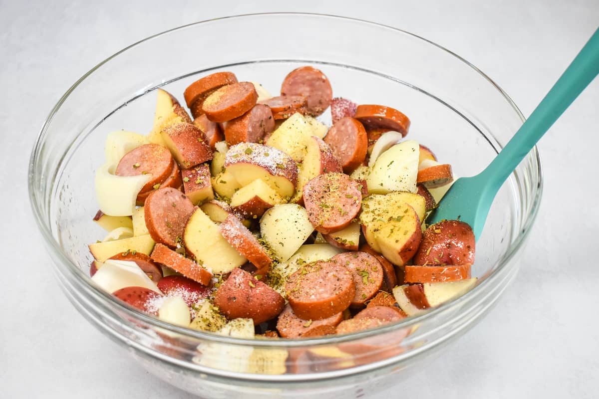 Seasoning added to sausage, potatoes, and onions in a large, glass bowl, with a teal colored silicone spatula on the right side.