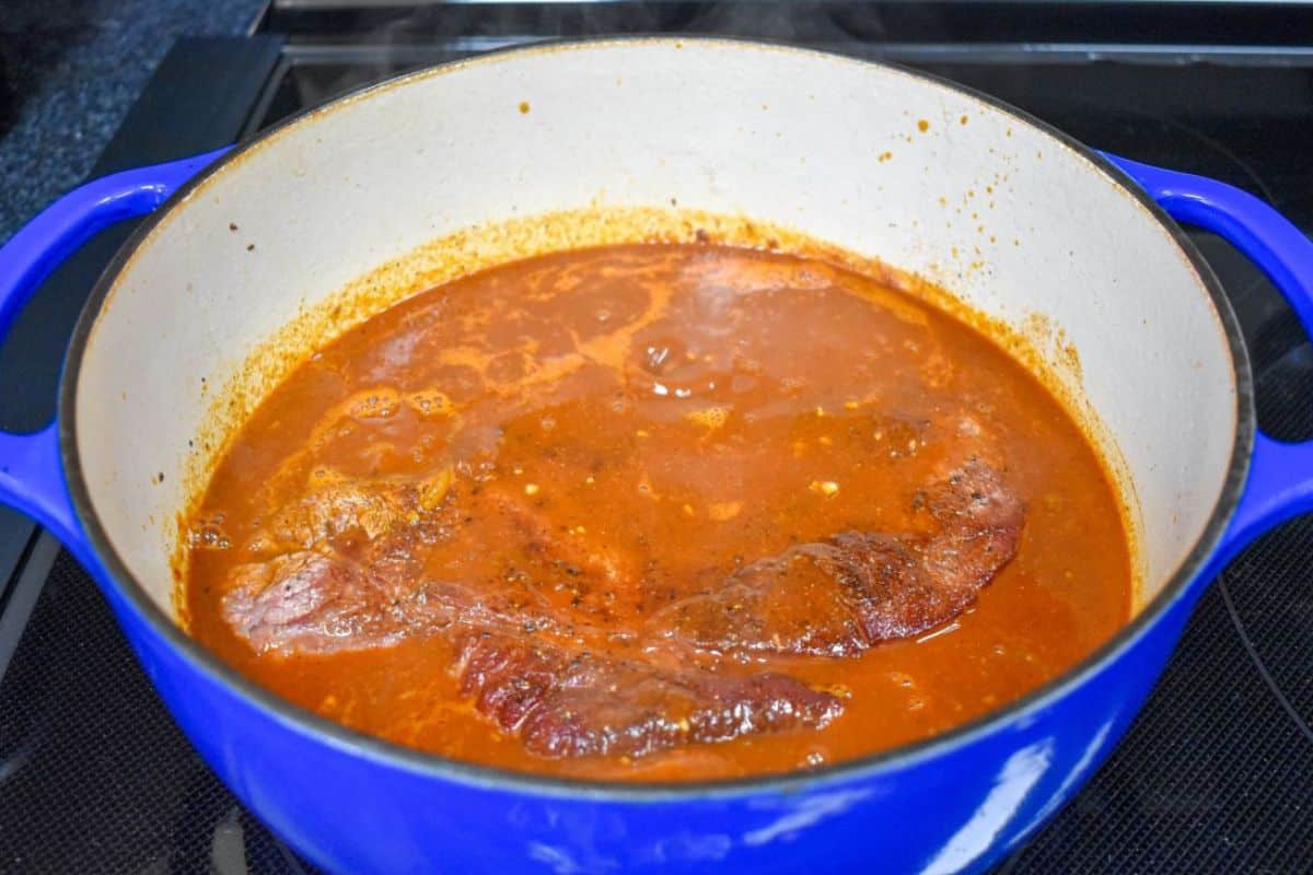 Browned beef chuck roast simmering in the tomato-based sauce in a Dutch oven.