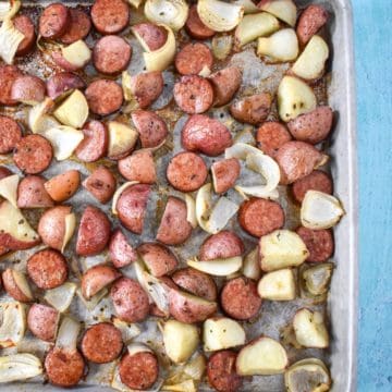 The finished sausage and potatoes on a sheet pan set on a light blue table.
