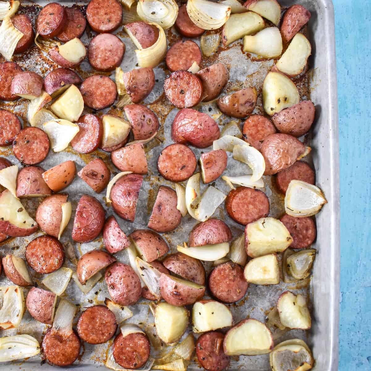 The finished sausage and potatoes on a sheet pan set on a light blue table.