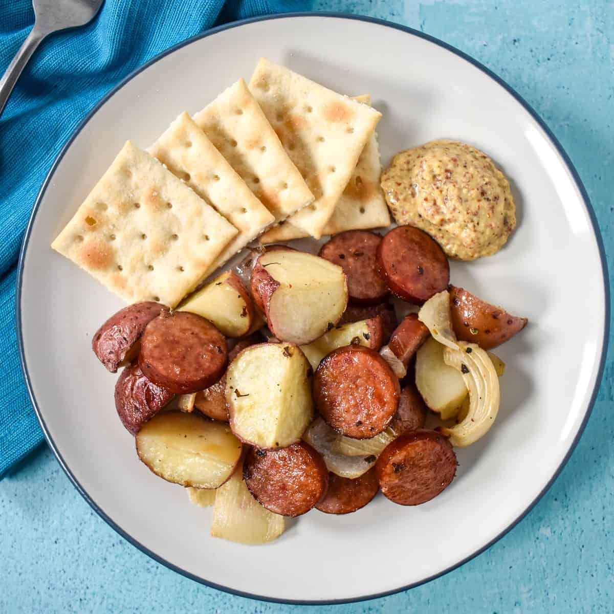 The sausage and potatoes served with brown mustard and saltine crackers on a white plate set on an aqua colored table with a teal linen.