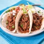 Three shredded beef tacos topped with cilantro and diced onion, served with lettuce, tomatoes, and lime.