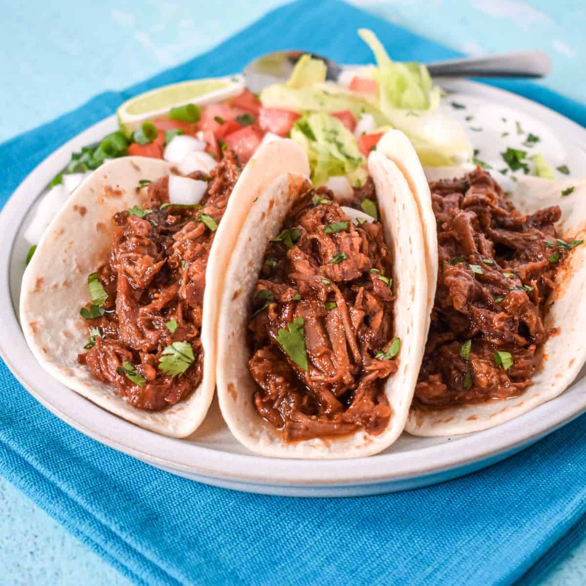 Three shredded beef tacos topped with cilantro and diced onion, served with lettuce, tomatoes, and lime.