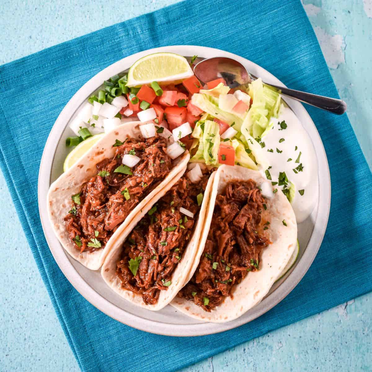 Overhead view of three shredded beef tacos with cilantro and onion, served with lettuce, tomatoes, lime, and sour cream.