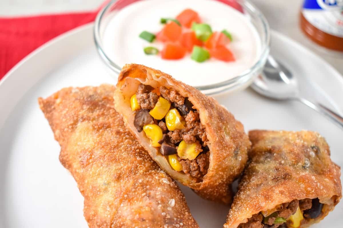 Two egg rolls served on a white plate with sour cream garnished with diced tomatoes and green onions.
