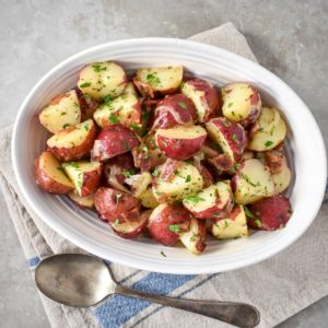 The warm potato salad served in an oval, white bowl with a large serving spoon on the side.