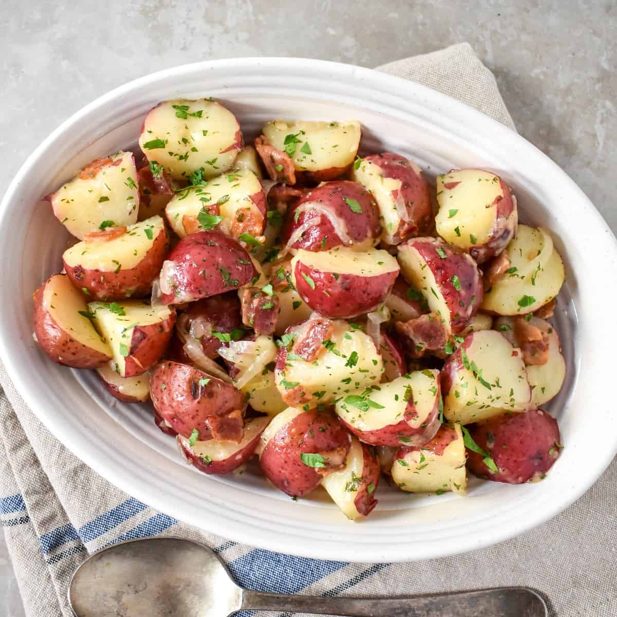 The warm potato salad served in an oval, white bowl with a large serving spoon on the side.