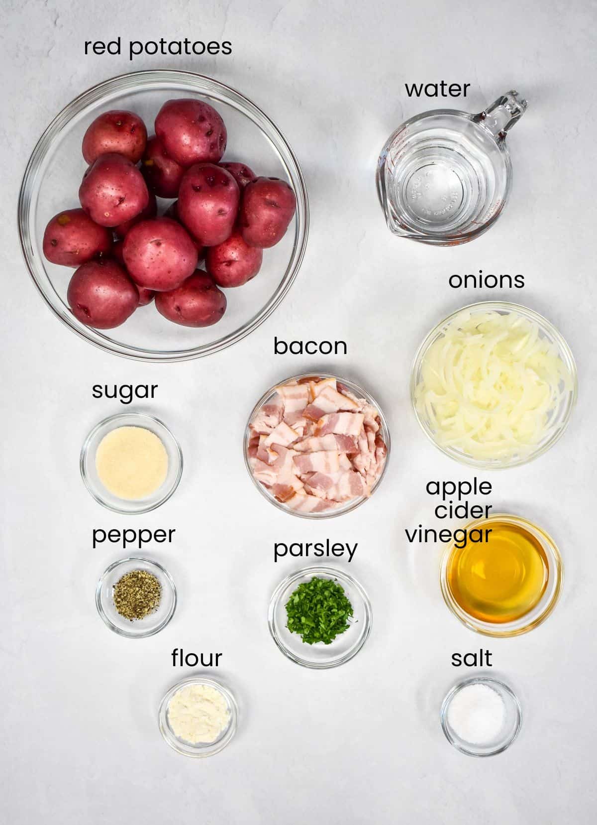 The ingredients for the warm potato salad arranged in glass bowls on a white table. Each ingredient is labeled with small black letters.