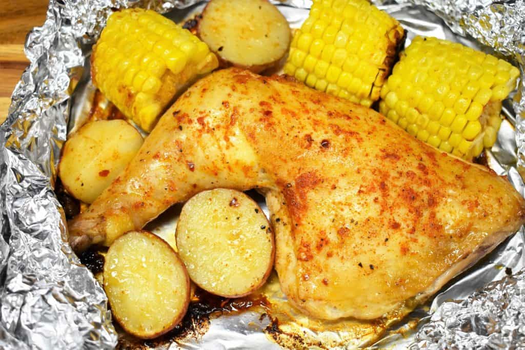An open foil pack with a quarter chicken, corncobs and potatoes cut in half.
