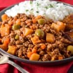 The picadillo served with white rice and fried sweet plantains on a gray plate on top of a red linen.