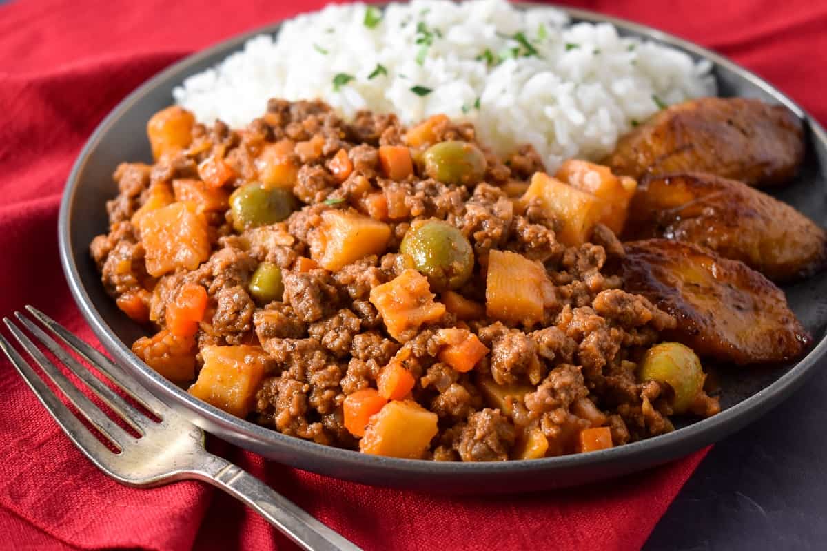 The picadillo served with white rice and fried sweet plantains on a gray plate on top of a red linen.